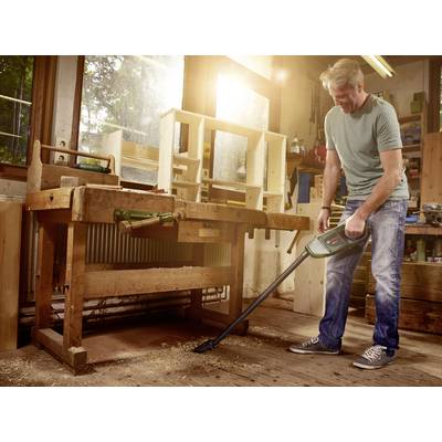 A man is hoovering wood shavings in a workshop. A workbench and wooden shelves are in the background. Sunlight streams through the window.