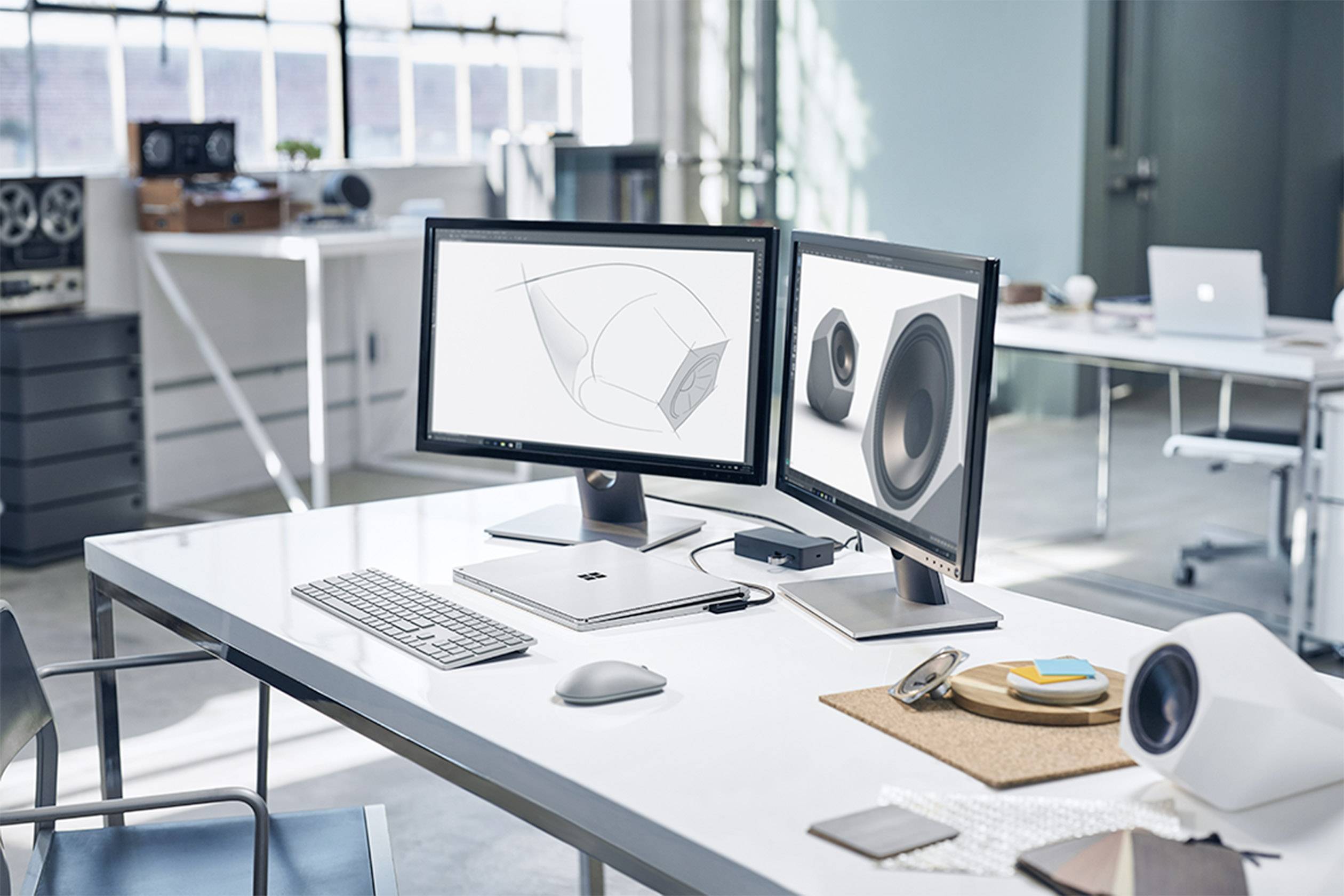 Modern office with two monitors displaying 3D designs. A keyboard, mouse, and speakers sit on the desk. Bright, minimalist interior.