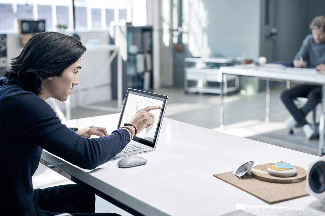A man is sitting at a desk, working with a digital pen on a laptop. An office is visible in the background.