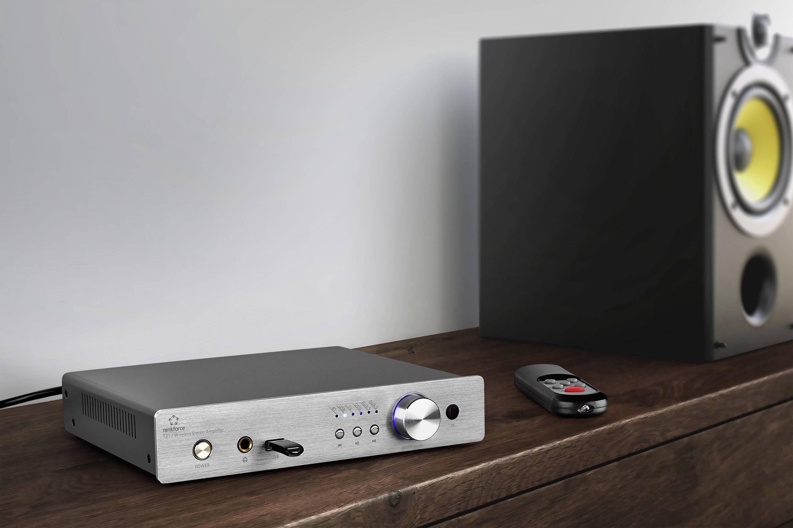 On a wooden table sit a compact amplifier with controls and displays, a remote control, and a modern speaker.