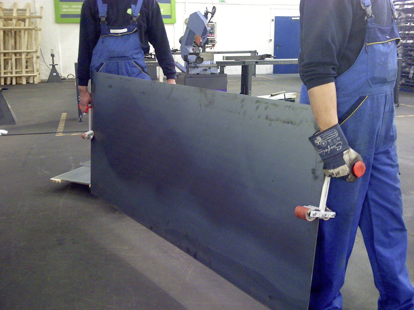 Two individuals wearing blue workwear are jointly carrying a large metal sheet using special clamps in a workshop environment.