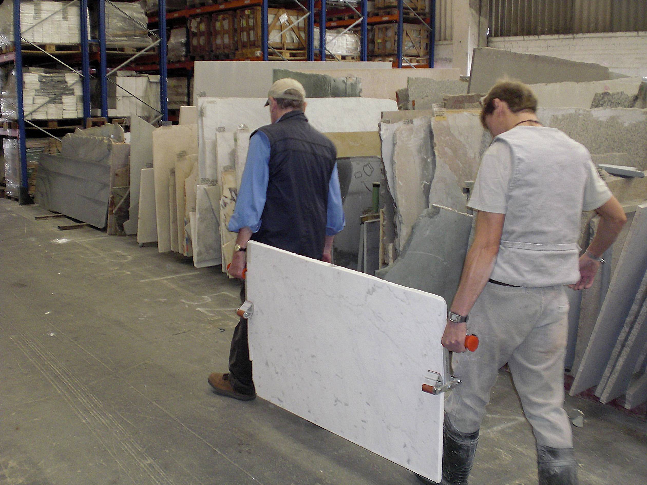 Two men are carrying a white marble slab through a warehouse filled with stone slabs. The floor is grey and the warehouse has tall shelves.