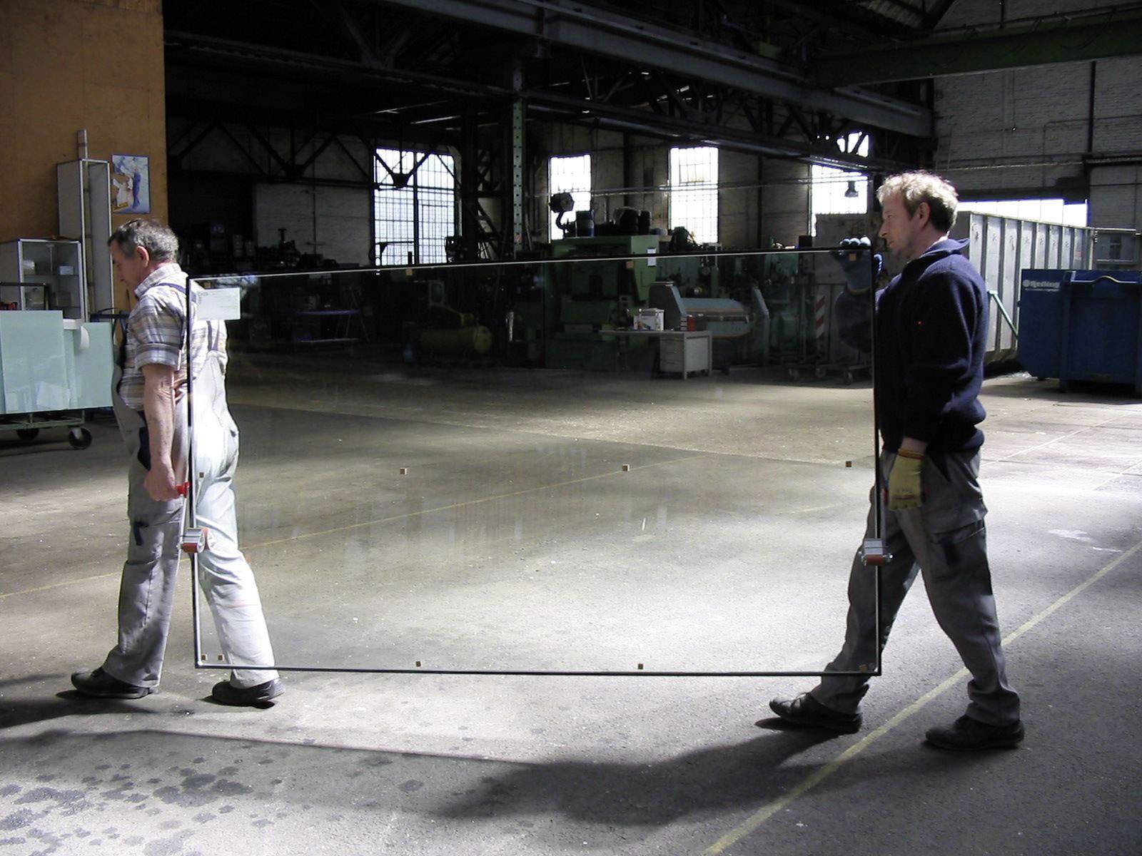 Two people are carrying a large glass pane in an industrial hall. The hall is dimly lit and appears spacious.