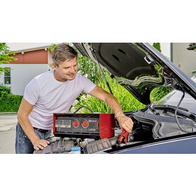 A man is jump-starting a car with a battery booster outdoors. The bonnet is open; the man is wearing a white T-shirt.