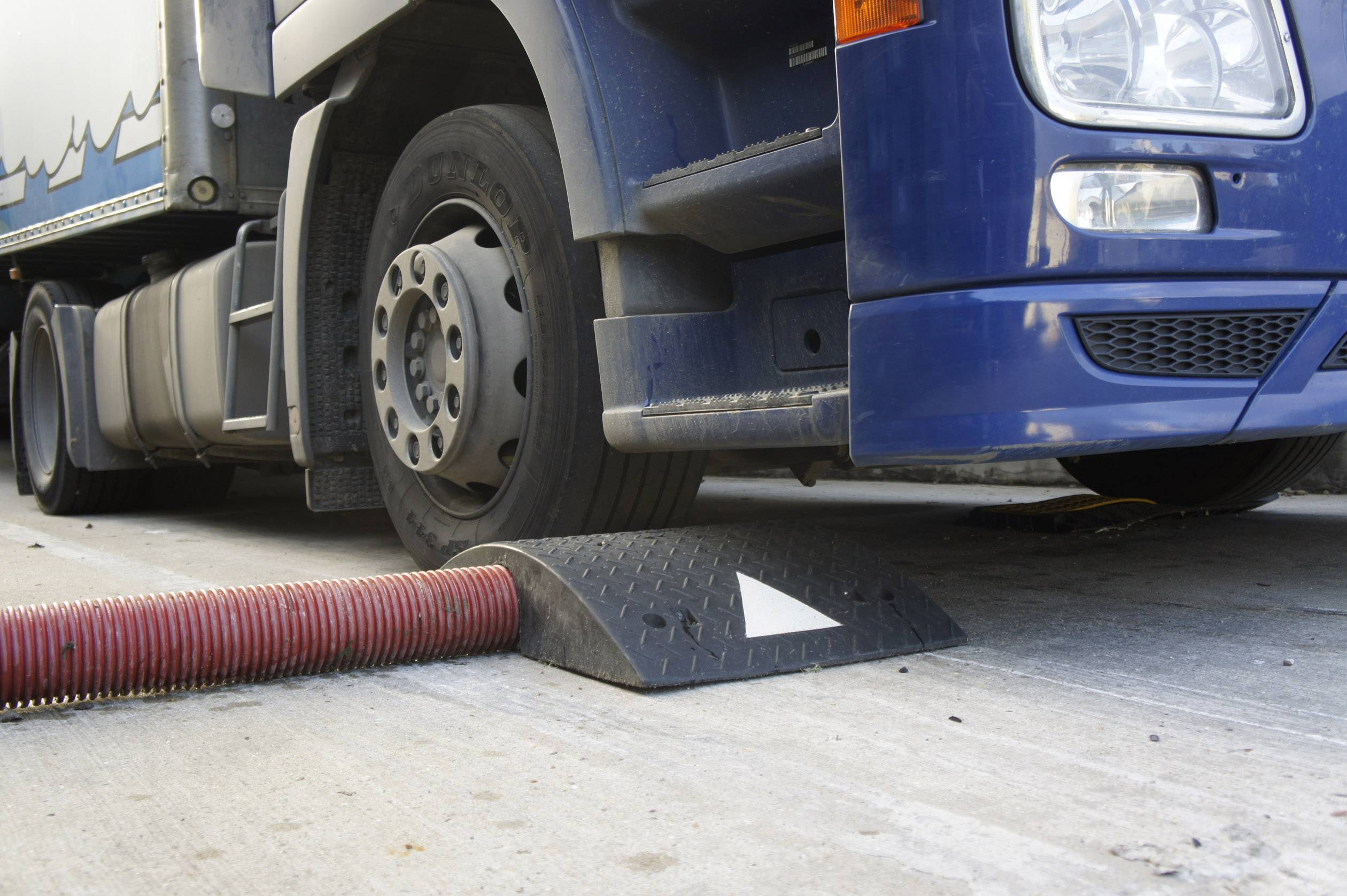 Lorry tyres are partially positioned on a black ramp with a white arrow, next to a red hose on a concrete structure.