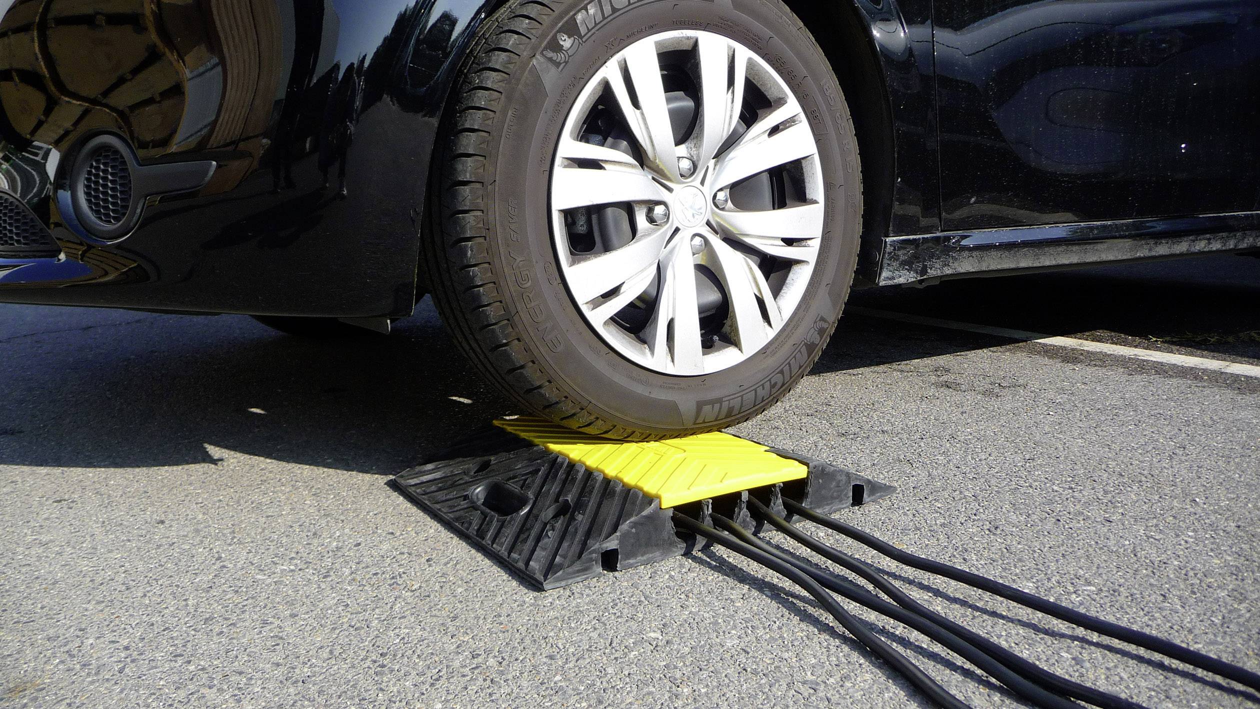 A car tyre drives over a yellow, diamond-shaped cable bridge element on a car park, which protects several cables.