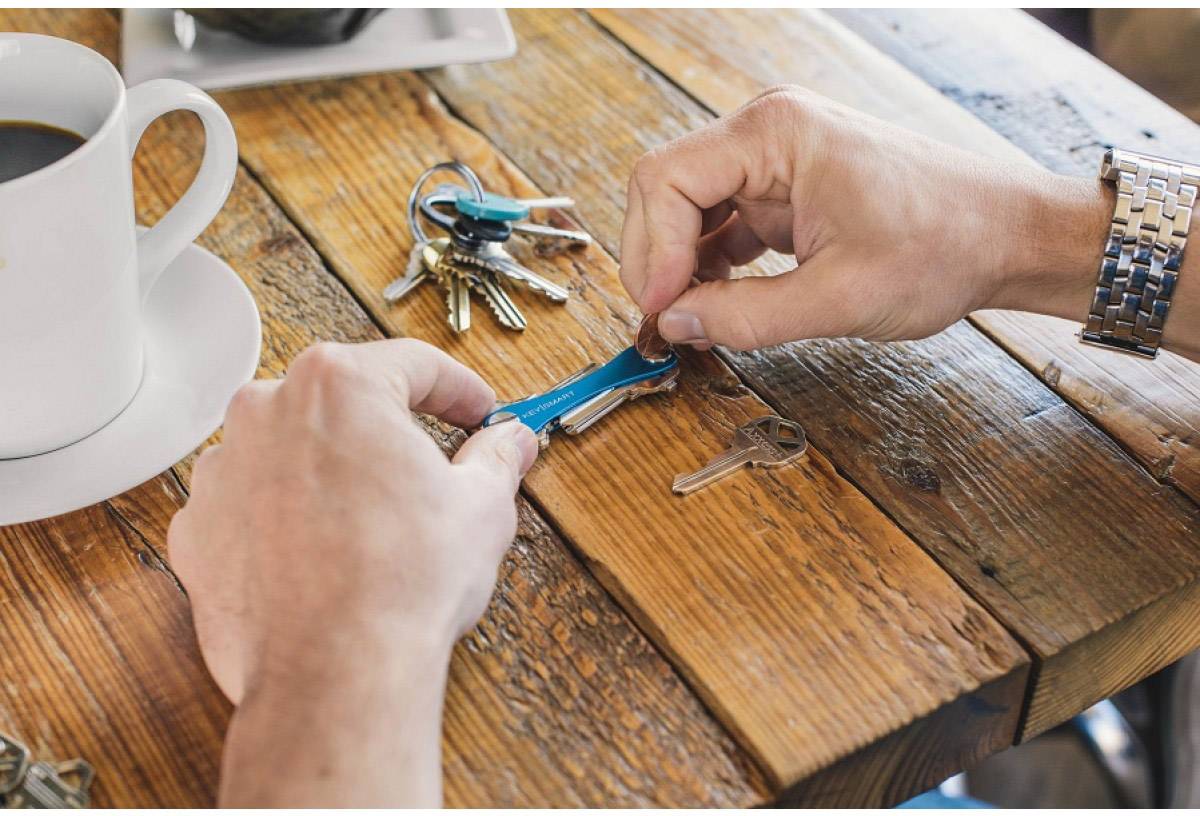 A person is attaching a key to a blue keyring on a wooden table, with additional keys and a cup of coffee nearby.