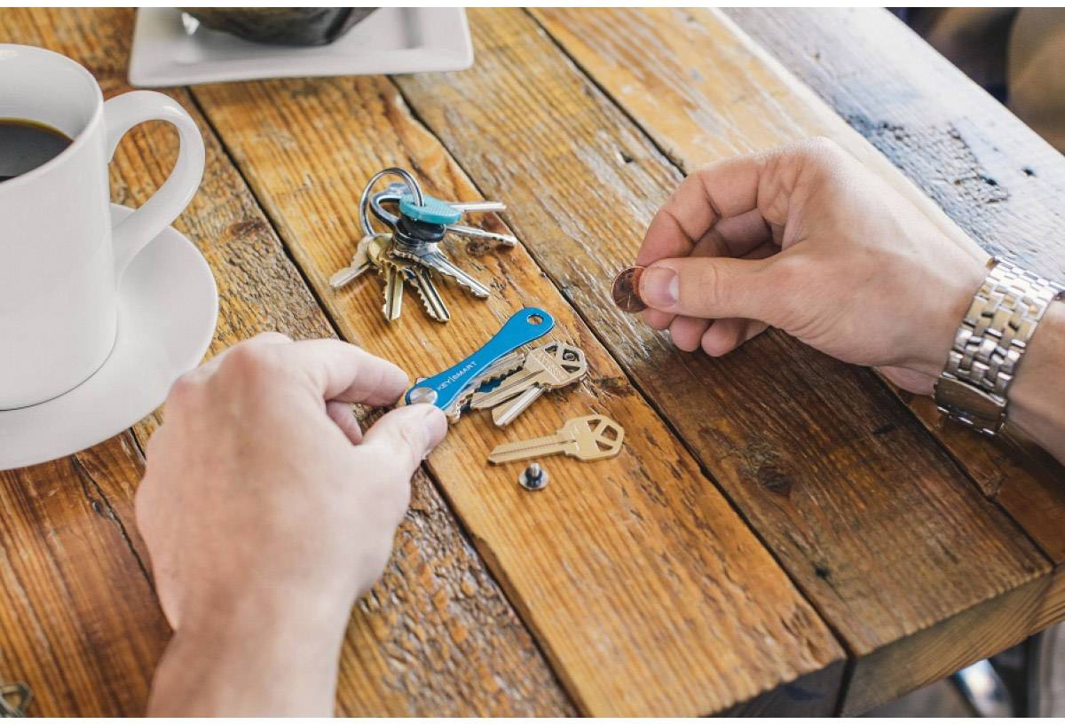 A person is sorting keys on a wooden table, next to a cup of coffee. They are holding a coin in their hand.