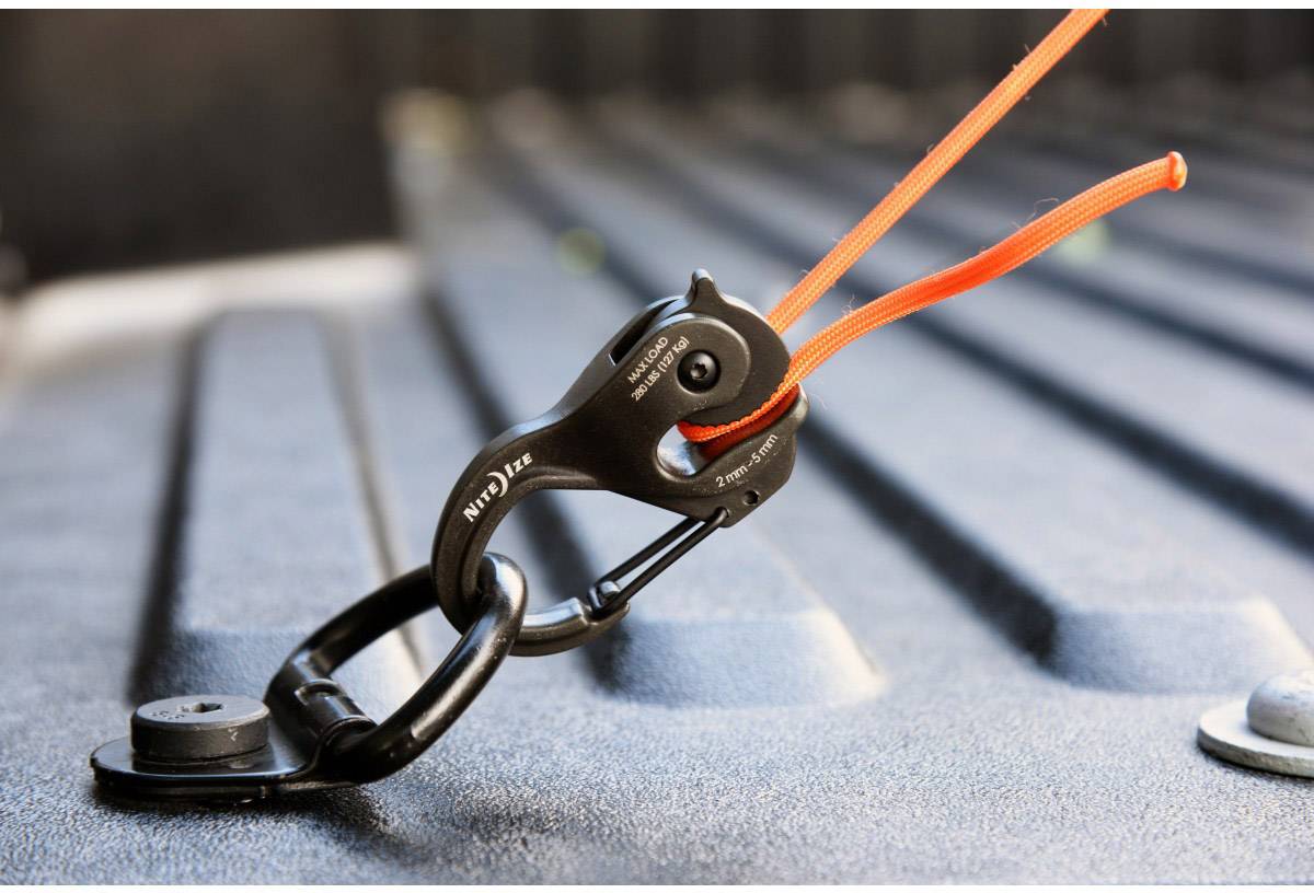 A black carabiner secures an orange rope on a metallic surface, possibly for cargo restraint.