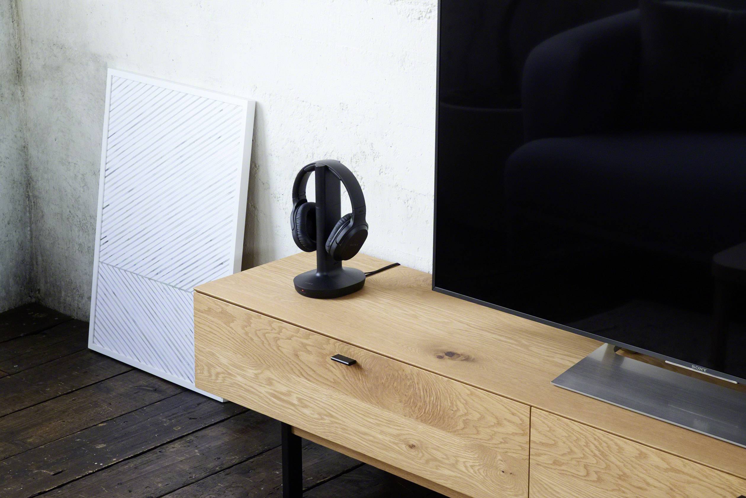 A modern living space with a wooden TV cabinet, on which black headphones are placed next to a flat-screen television. A picture hangs on the wall in the background.