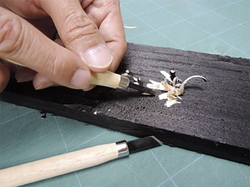 A hand carves with a knife on a dark piece of wood, wood shavings gathering. Another knife lies beside it on a green surface.