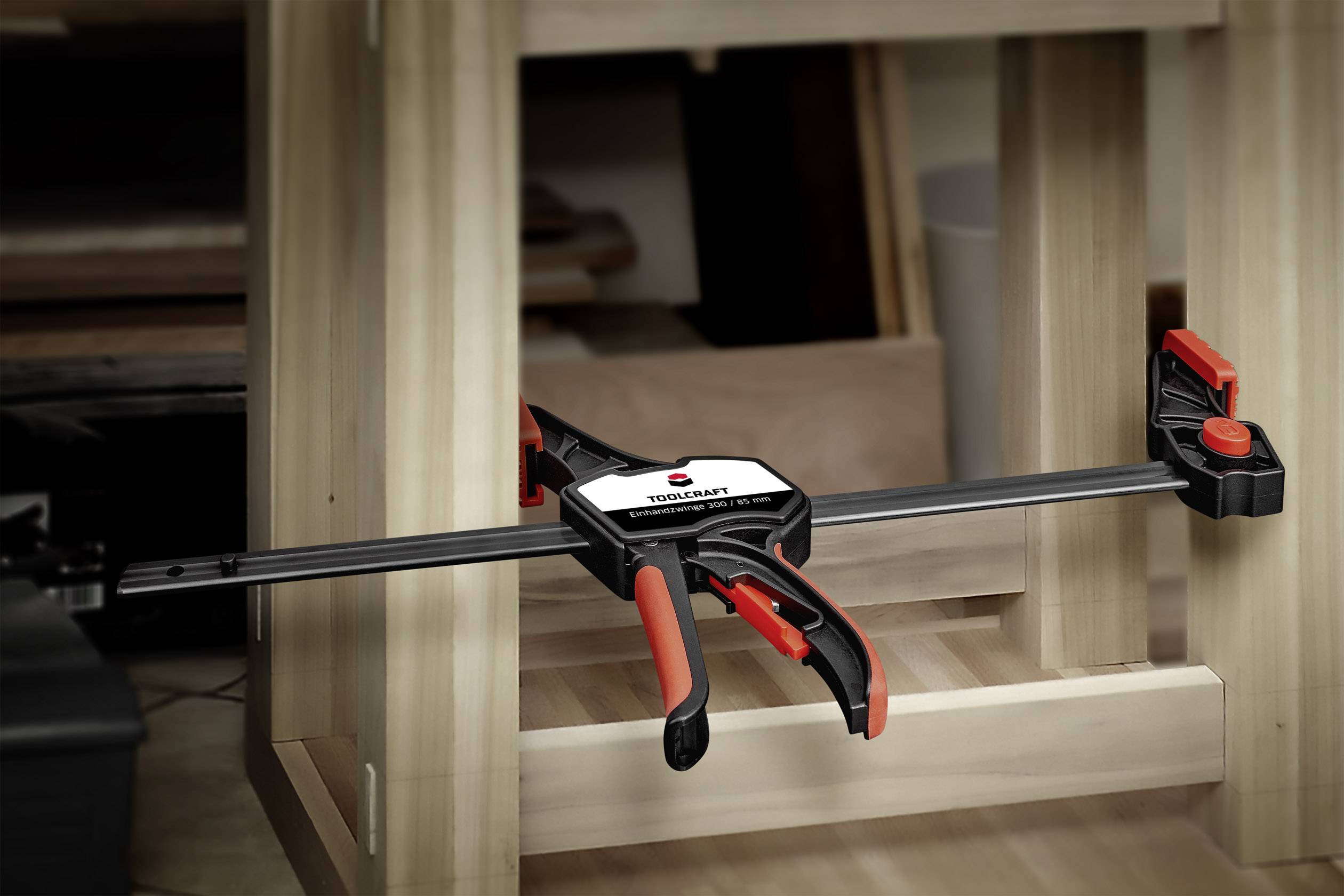 A black one-handed clamp with red handles securely grips a piece of wood to a wooden table.