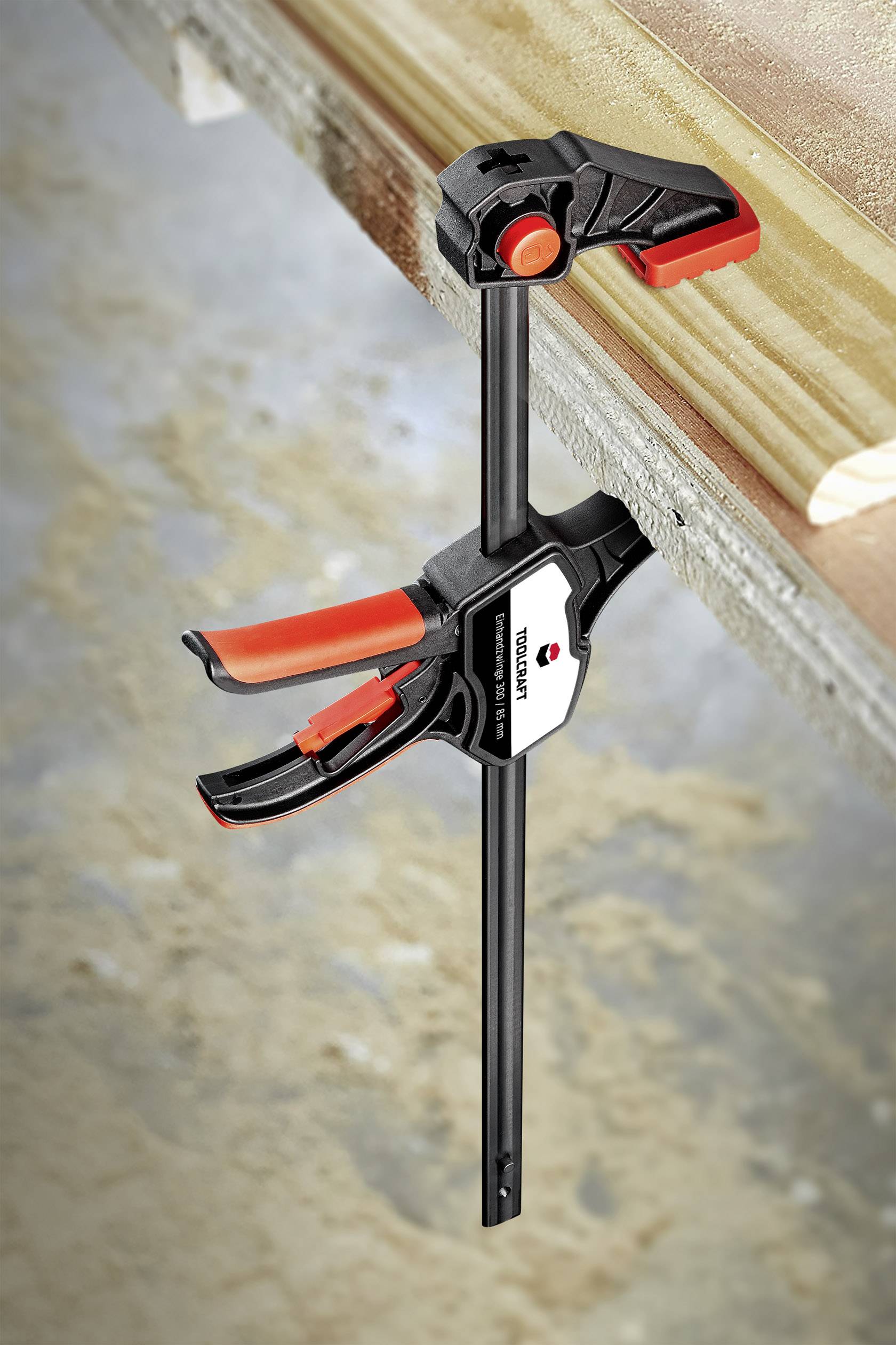 Woodworking Workshop: A clamp holds a plank firmly on a workbench. It has a red handle and a black frame.