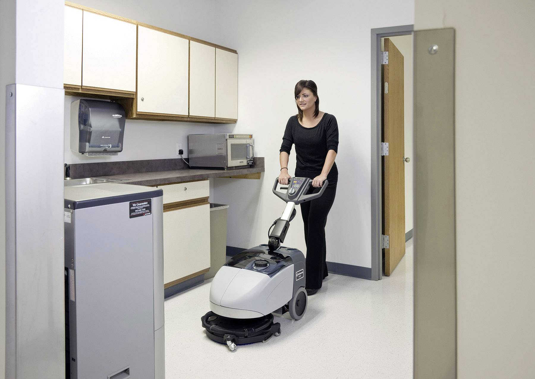 A woman is cleaning the floor of an office building kitchen area with an electric cleaning machine.