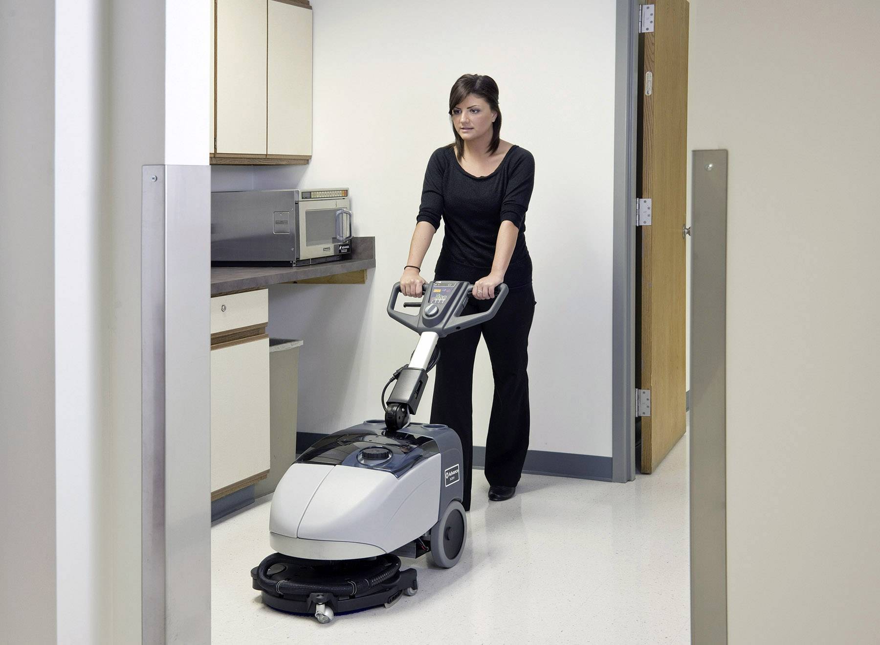 A person is using a floor scrubbing machine in an office corridor. Cupboards and a microwave can be seen in the background.