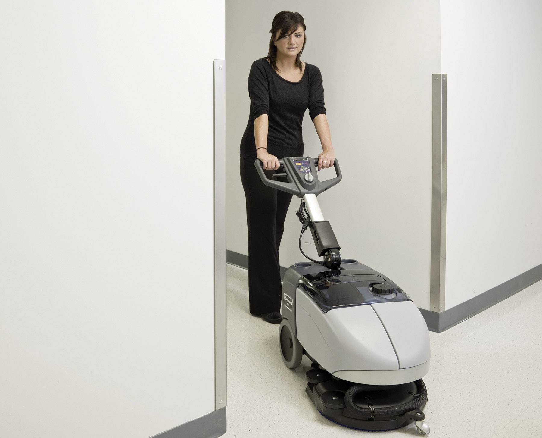 A woman is operating a modern floor cleaning machine in a corridor. She is wearing black clothing and working with concentration.
