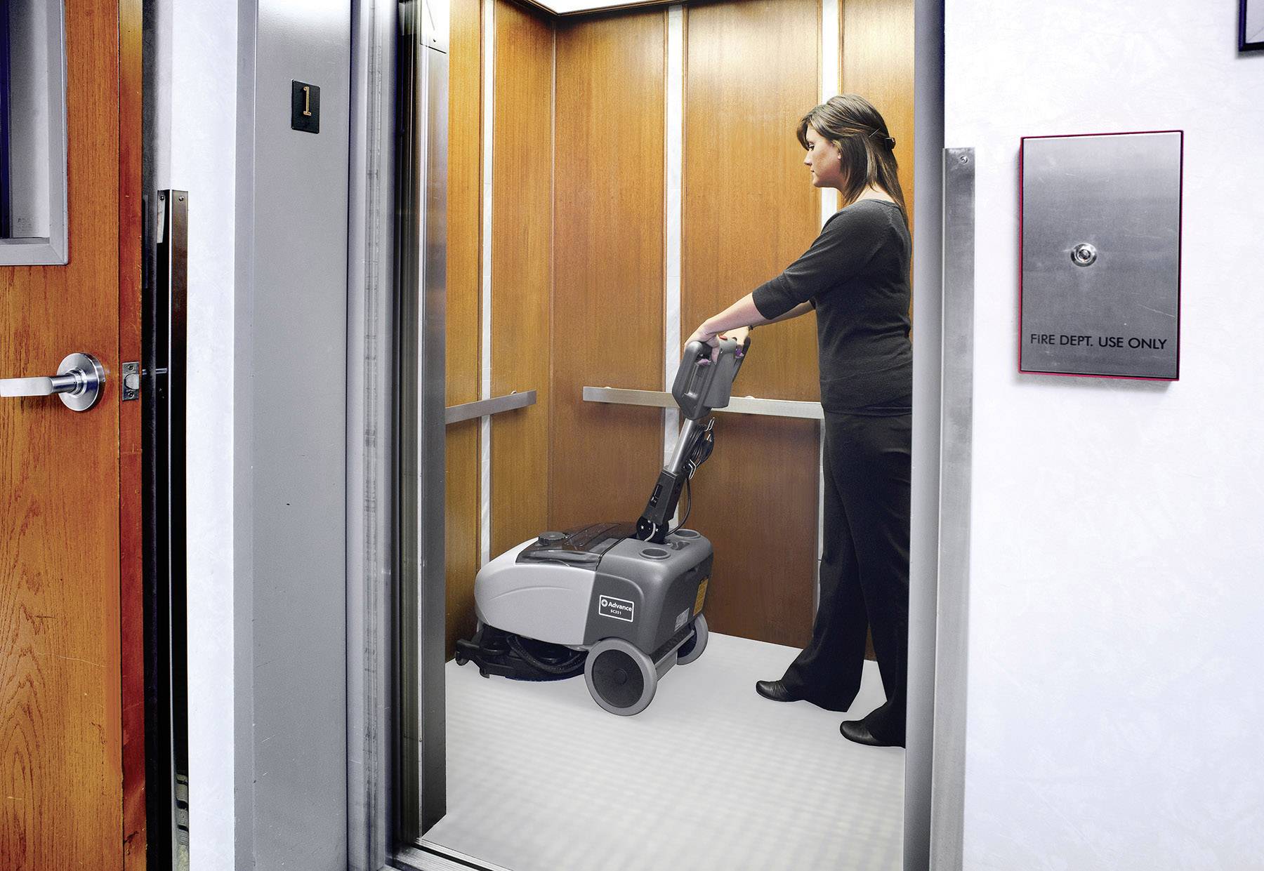 A person is cleaning the floor of a lift with a floor cleaning machine. The lift has wood-panelled walls.