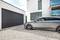 Silver car parked in front of a modern house and garage on a gravel area. Driver gazing out of the window towards the garage.