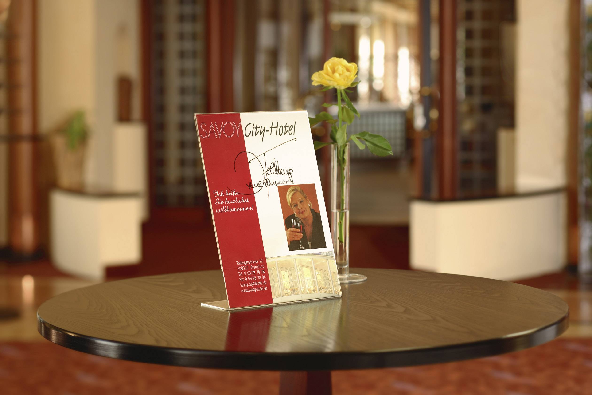 A round wooden table with a Savoy City Hotel sign and a yellow rose in a vase, situated in an elegant hotel lobby.