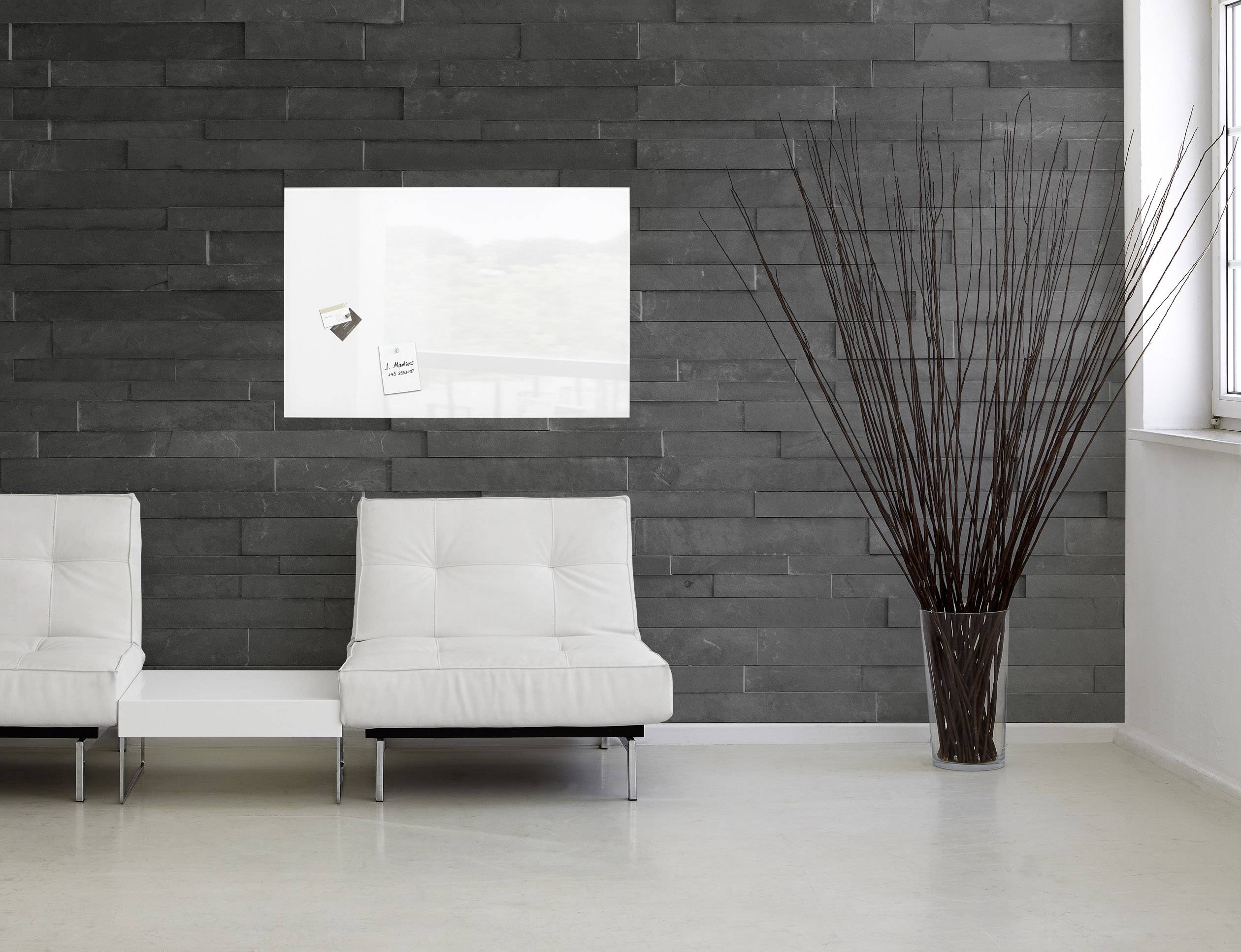 A bright room with two white armchairs in front of a dark stone wall. A rectangular artwork and a large floor vase with branches are visible.