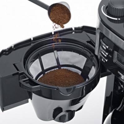 A coffee machine is being filled with coffee powder. A measuring spoon is pouring ground coffee into a filter in the open coffee machine compartment.