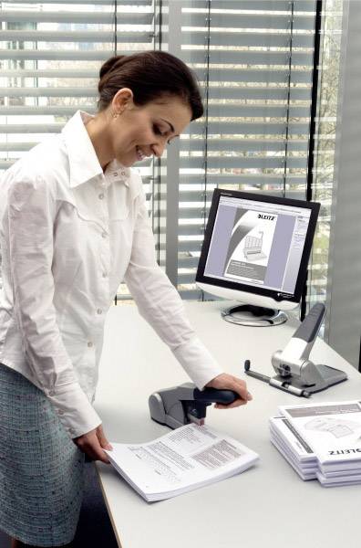 A woman in the office is scanning documents at a scanner. A computer screen displays an open document. Window blinds are visible in the background.