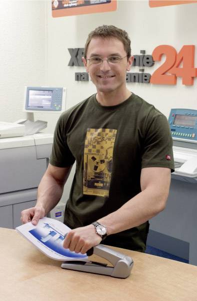 A person is standing smiling by a printer in an office, holding a document. Printing machines are visible in the background.