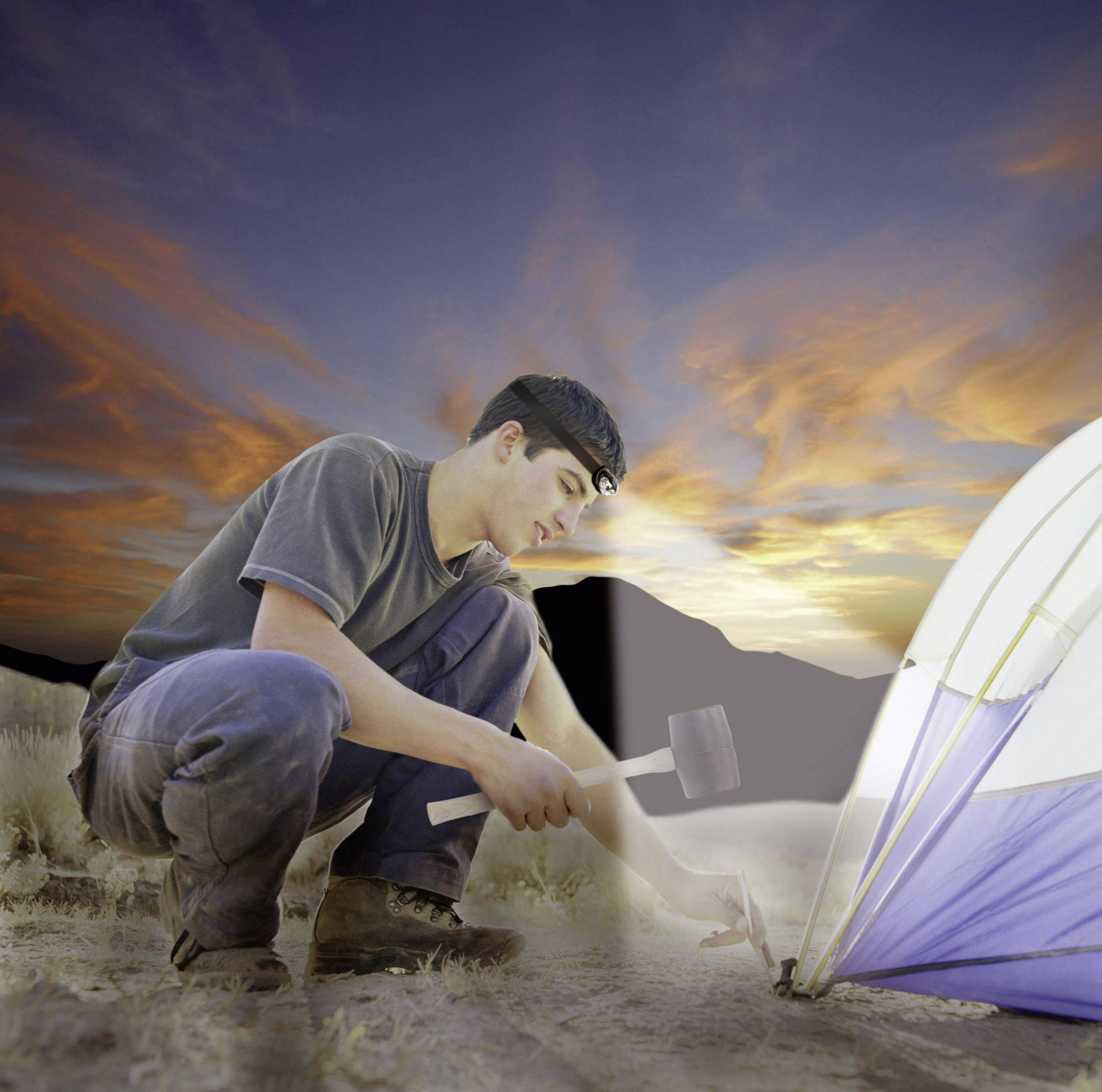 A man hammers a tent peg into the ground at sunset while setting up a tent outdoors.