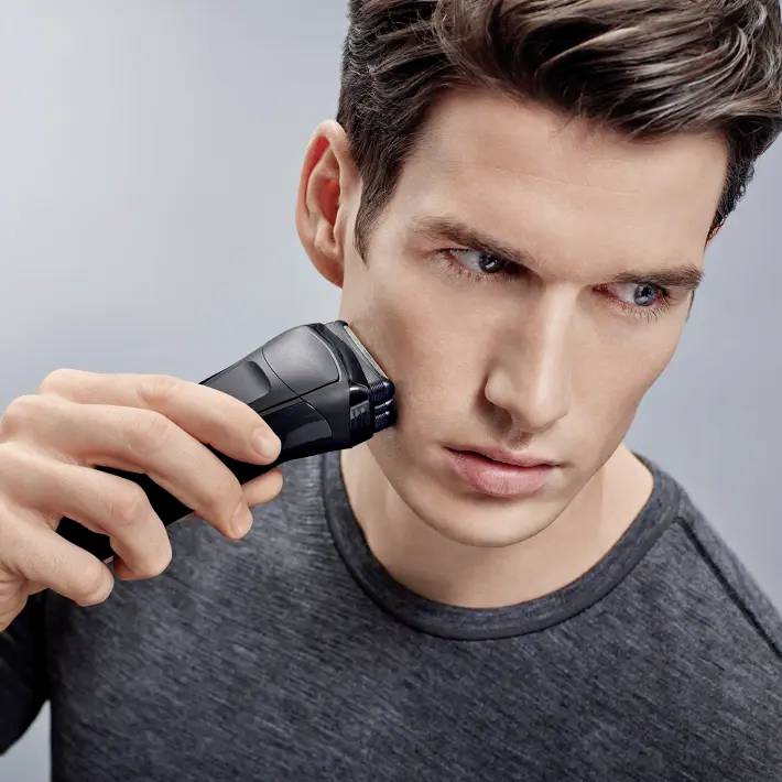 A man is shaving with an electric razor. He is wearing a grey T-shirt and has a concentrated facial expression.