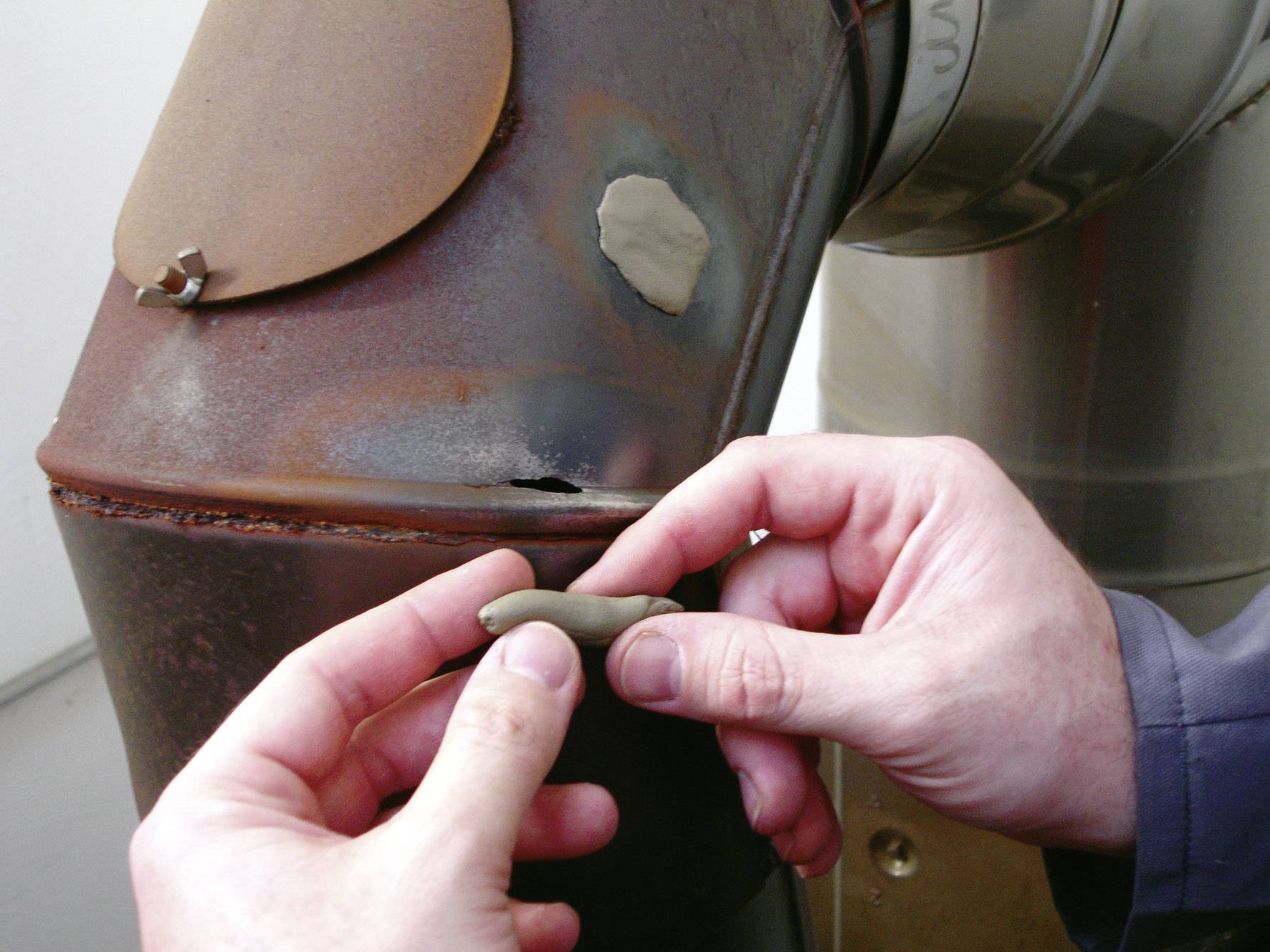 'Two hands are repairing a small hole in a rusty metal pipe using putty metal. The pipe is part of a larger metallic system.'
