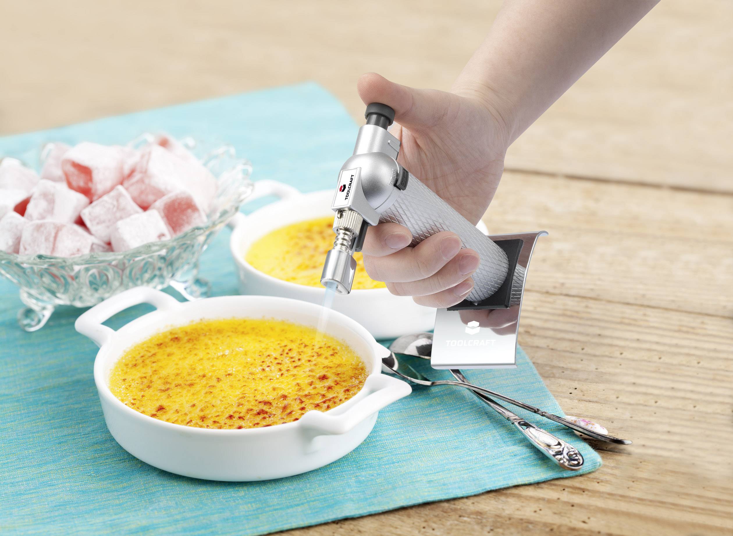 A hand holds a kitchen blowtorch to caramelise the sugar surface of crème brûlée in white ramekins. A glass dish of Turkish delight is on the right.