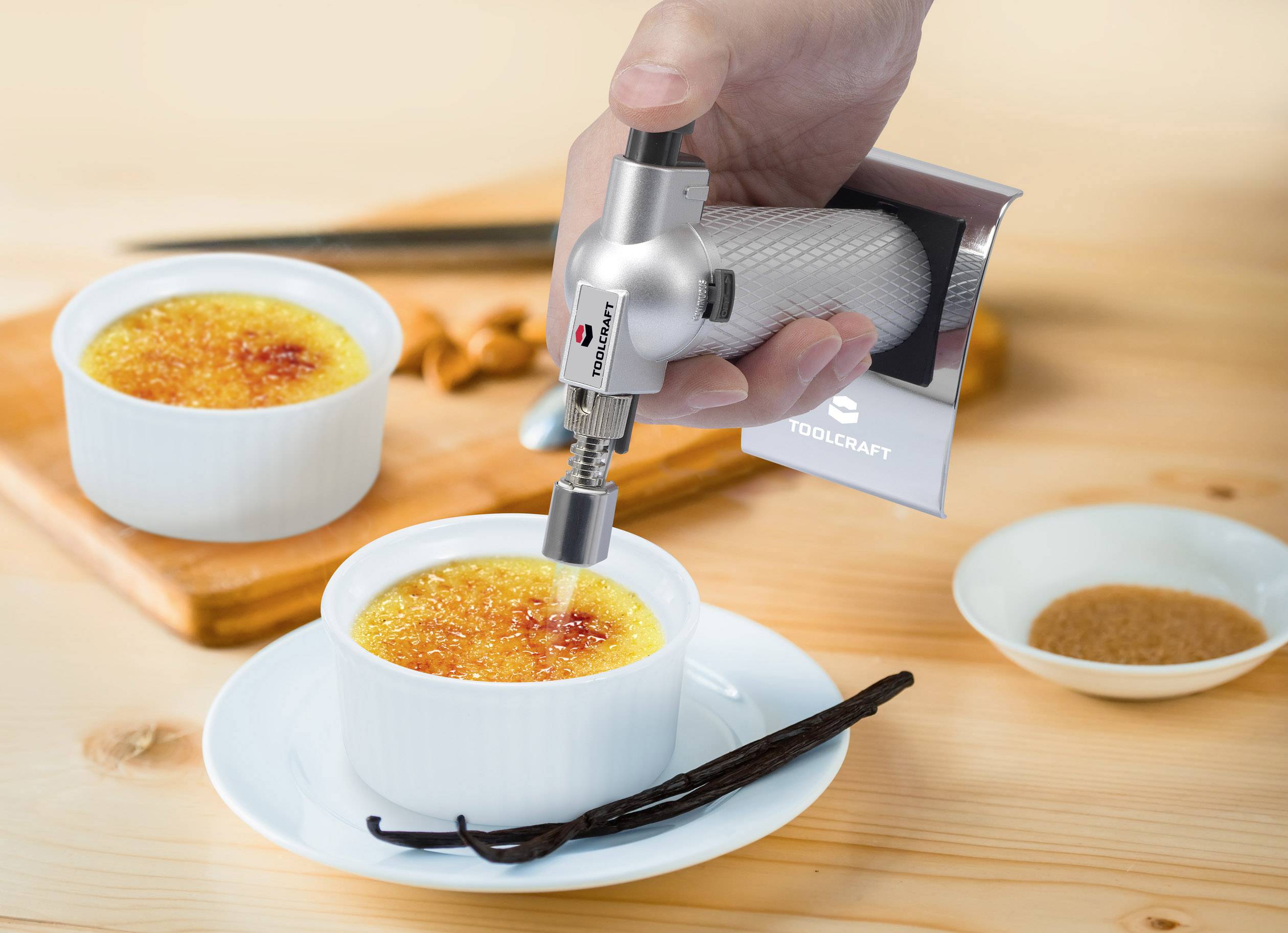 A hand burns sugar on a crème brûlée using a blowtorch. Two bowls on plates, with vanilla pods and a wooden board in the background.
