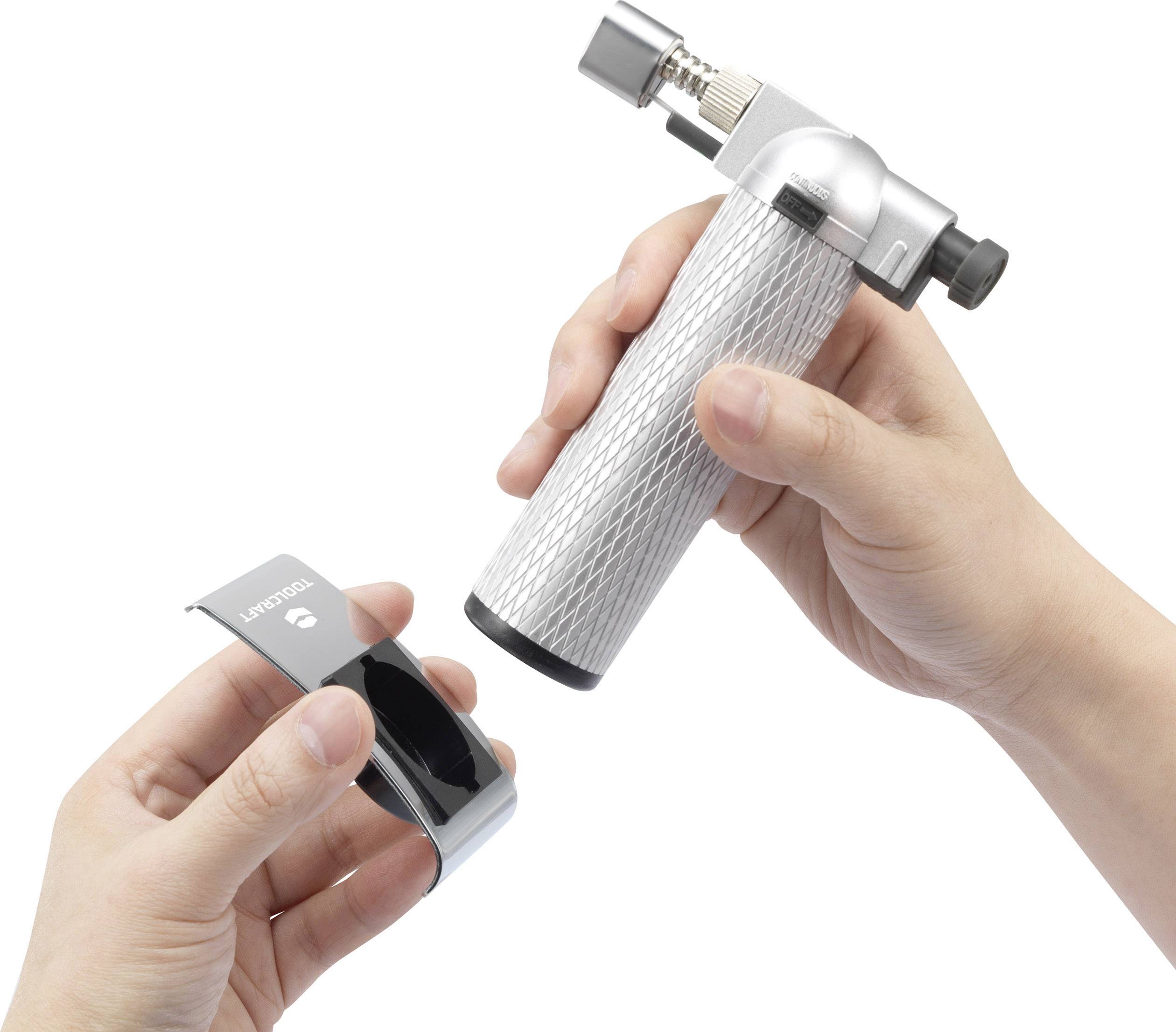 A person is holding a small, silver blowtorch in their right hand and a black, metallic stand in their left hand.