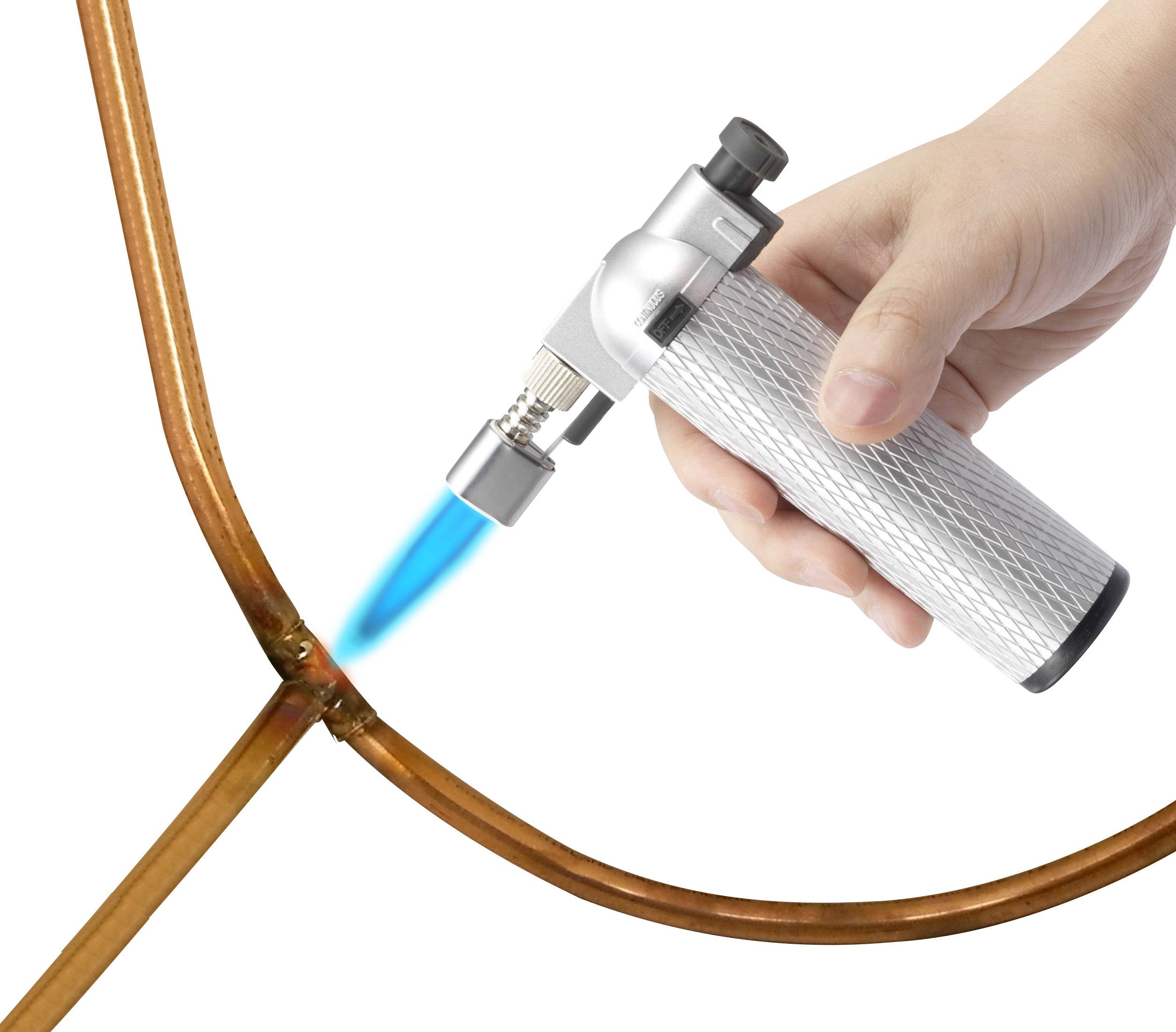 A hand holds a soldering torch and heats a copper pipe connection.