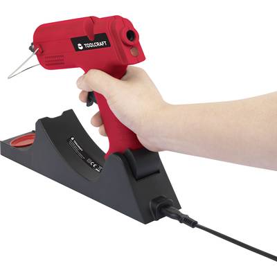 A red hot glue gun in a black charging station, held in a hand. It is designed for gluing small projects.