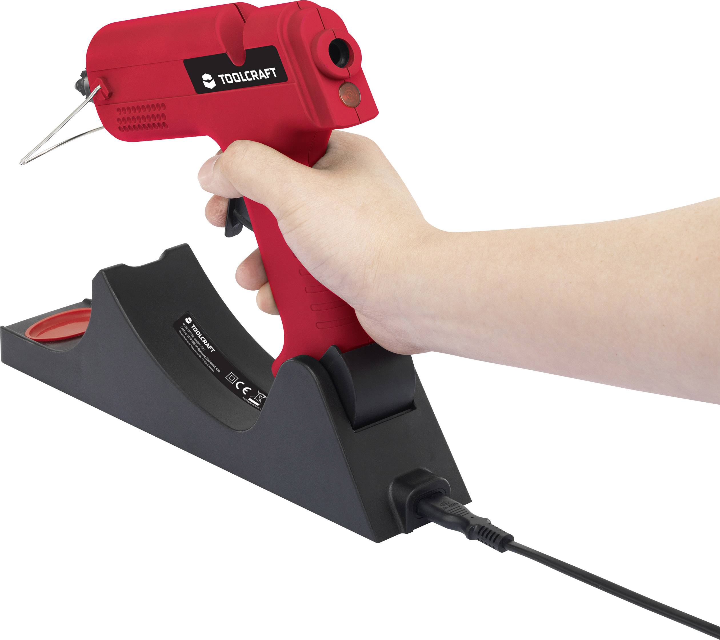 A hand is holding a red hot glue gun by Toolcraft, which is inserted into a black holder. A cable is connected to the holder.