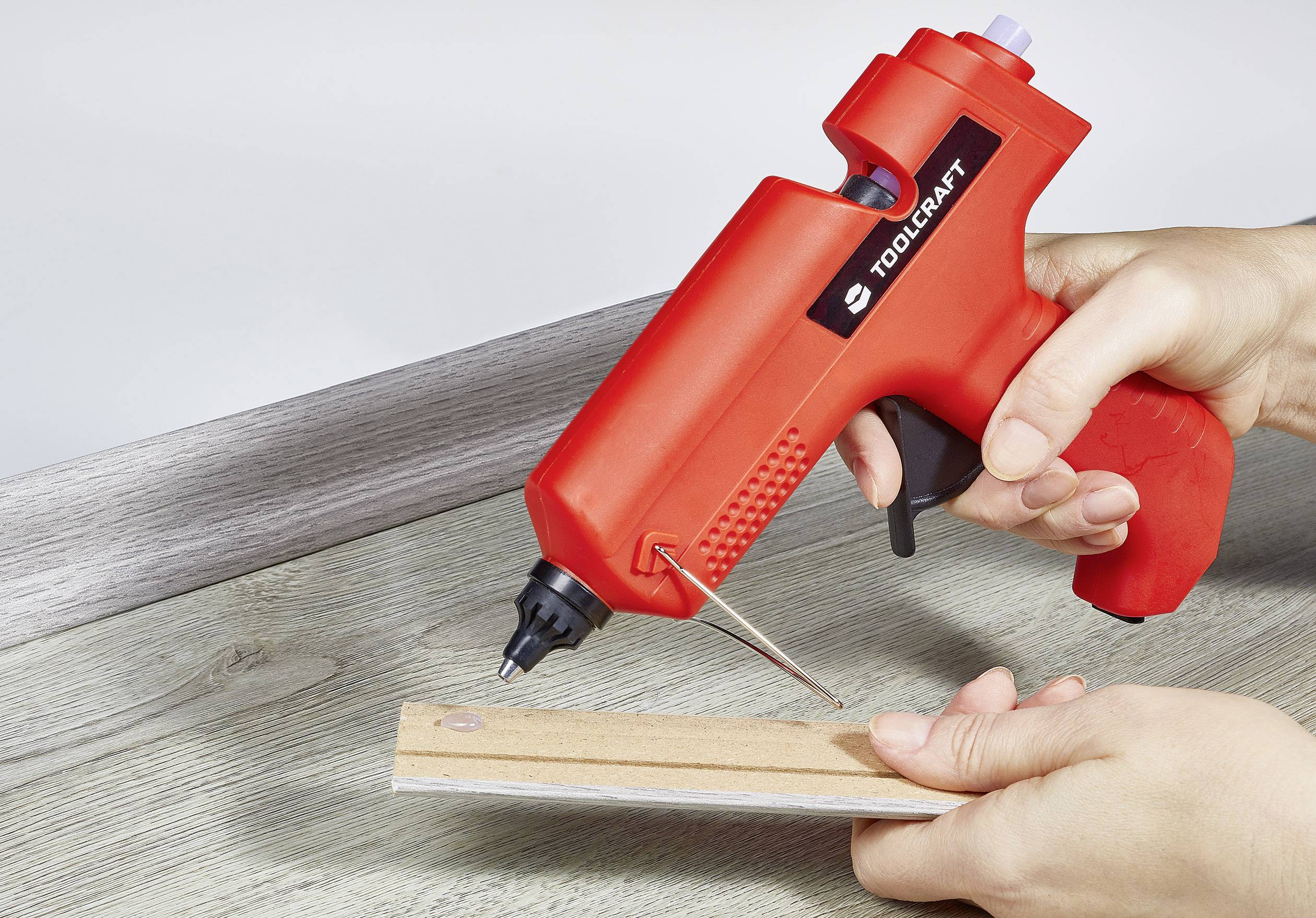 A person is using a red hot glue gun to stick a piece of wood onto a table surface.