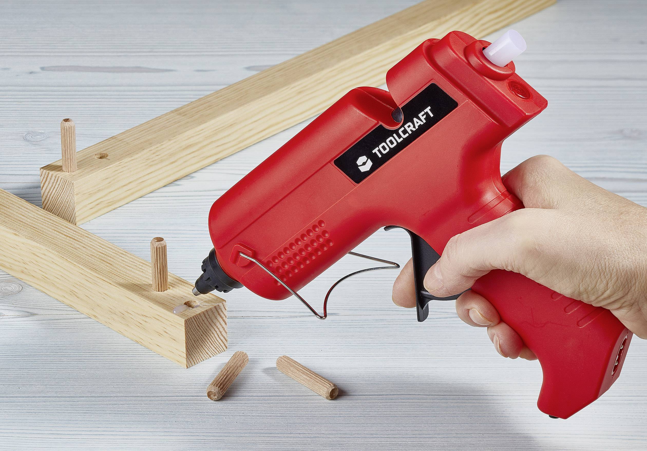 A hand holds a red hot glue gun while wooden dowels are being inserted into wooden pieces, likely for furniture assembly.