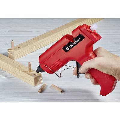 A hand holds a red hot glue gun while wooden dowels are being inserted into wooden pieces, likely for furniture assembly.