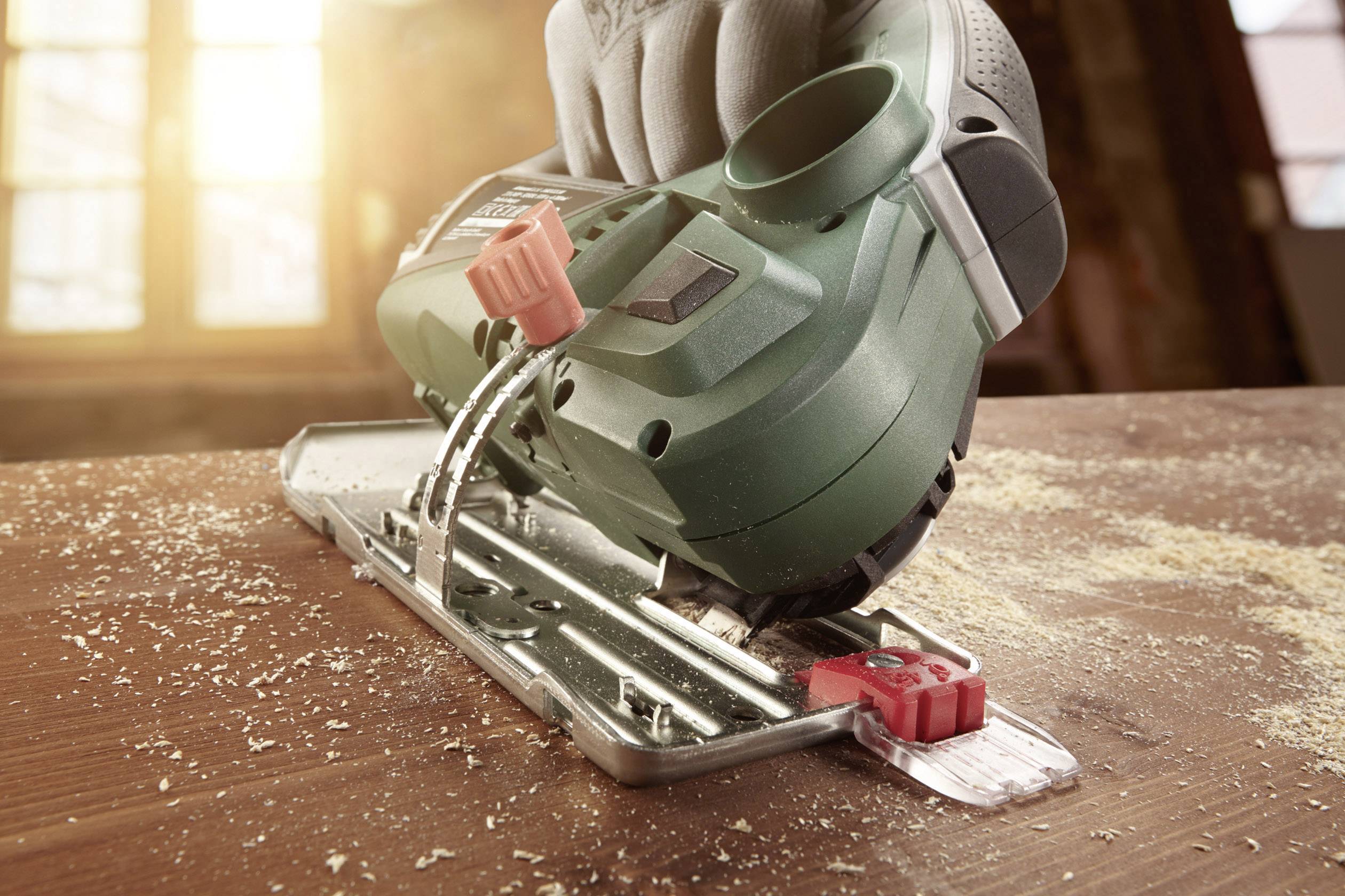 A hand holds an electric jigsaw gliding over a wooden board. Sawdust is dispersing around the cutting point.