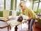 A woman is cutting a wooden board with an electric saw in a sunlit room. She is wearing safety glasses.