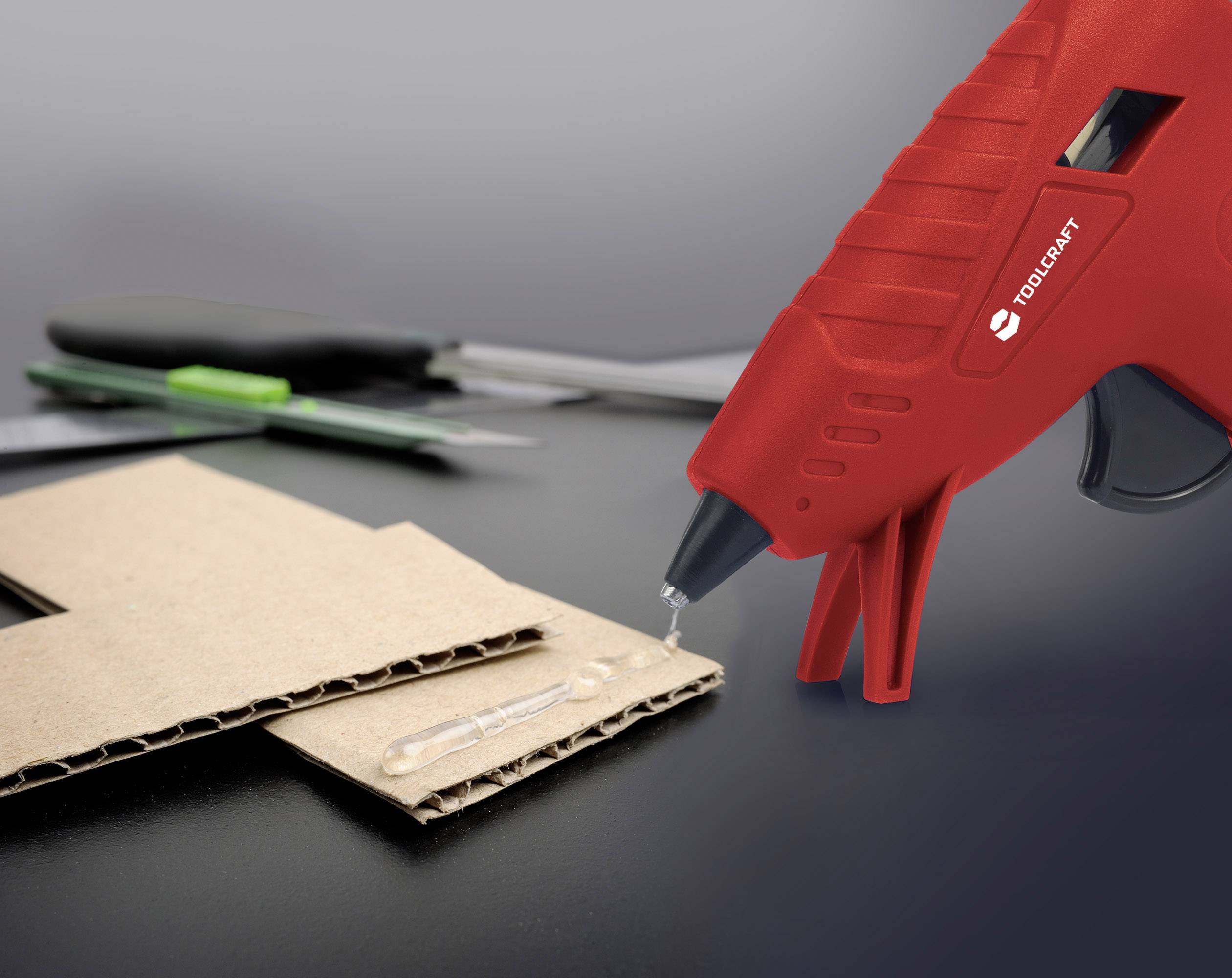 Red hot glue gun applying adhesive to a cardboard surface. A craft knife and another tool are lying in the background.