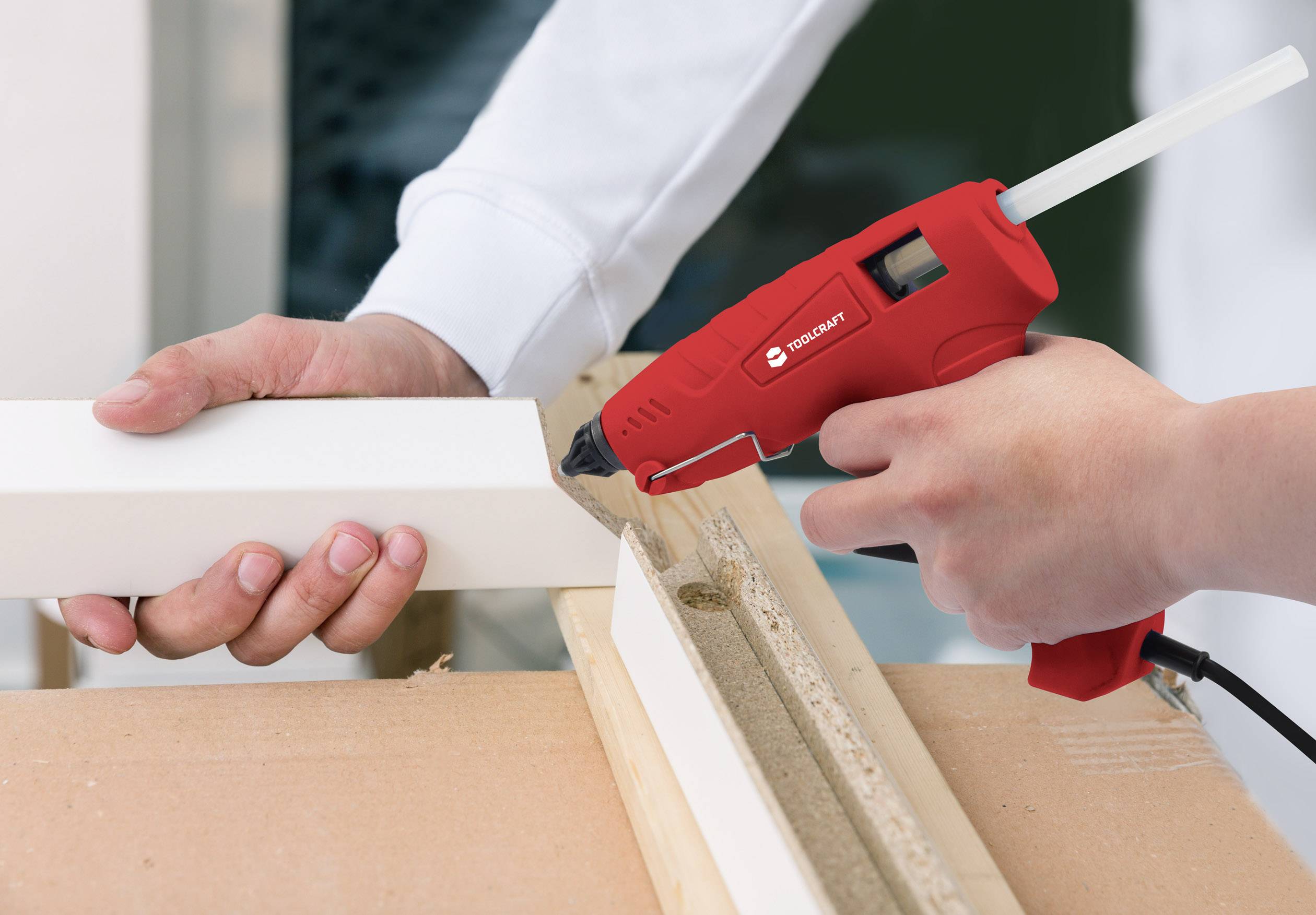 A person is using a red hot glue gun to adhere a white piece of wood. Hands are stabilising the workpiece on a wooden surface.