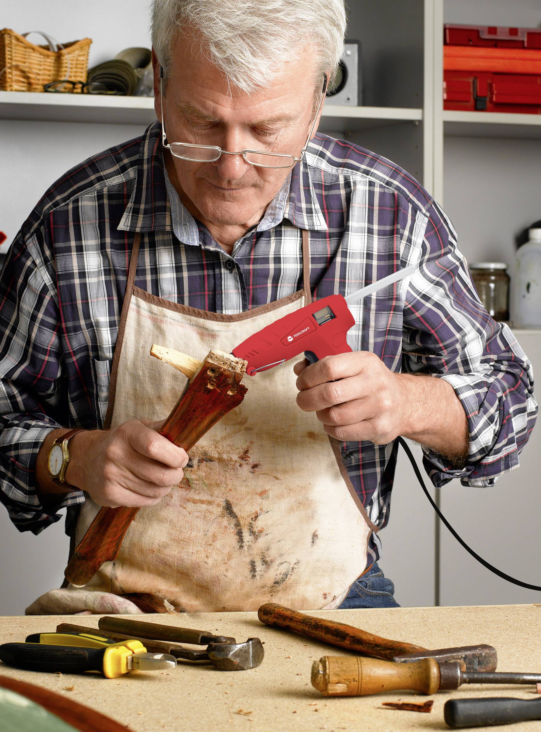 An elderly man is repairing a wooden leg with a red hot glue gun, surrounded by various tools in a workshop.