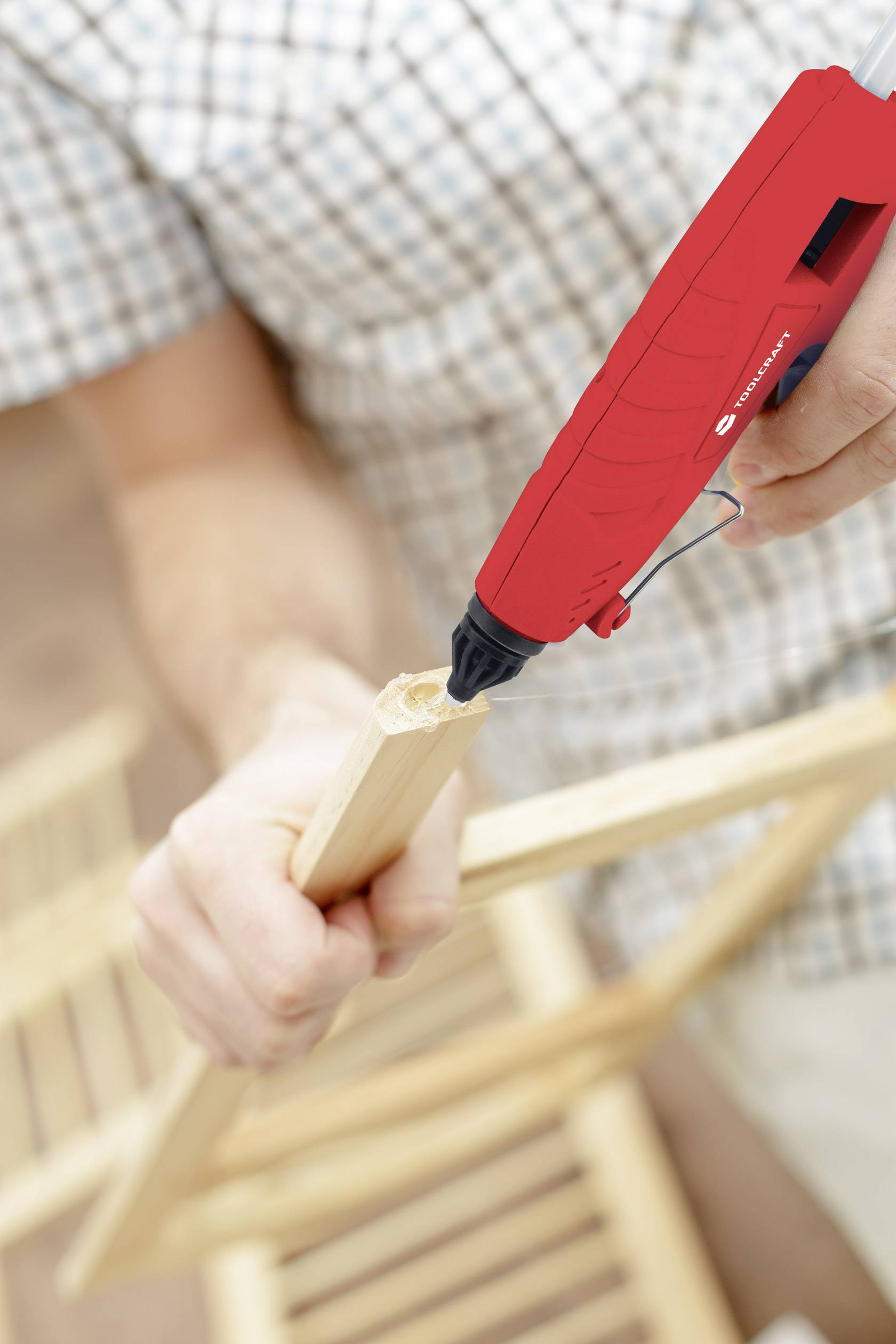 A person is wearing clothes with a checked pattern and using a red hot glue gun to stick wooden slats together. In the background, a wooden chair is visible.