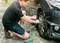 A man is changing a car tyre with a spanner, kneeling on a green cushion. The car is jacked up.