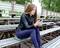 A woman sits on wooden benches outdoors and looks at her mobile phone. She is wearing a black leather jacket and dark trousers. The background shows trees.