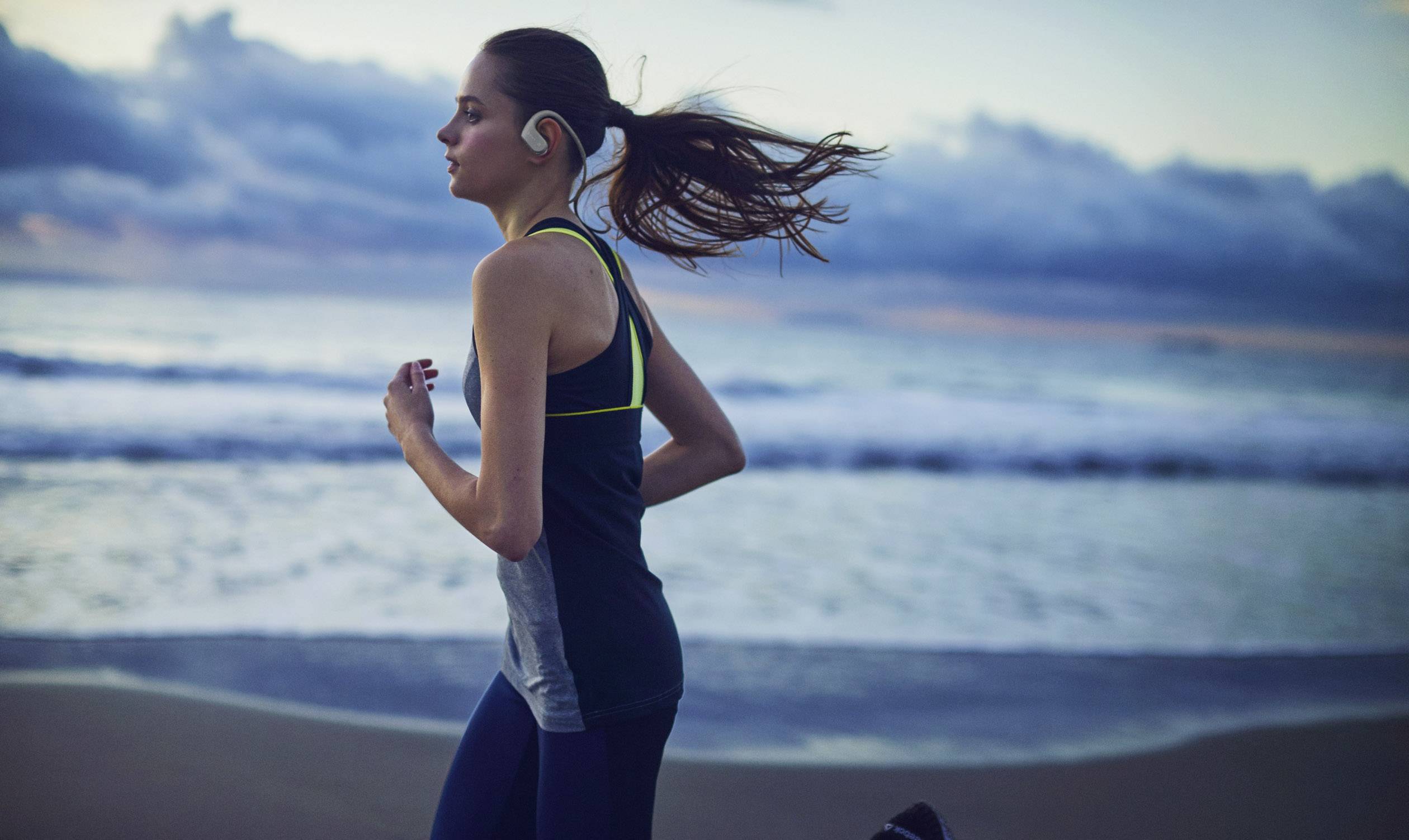 A woman jogs along the beach at sunset. She is wearing sportswear and headphones.