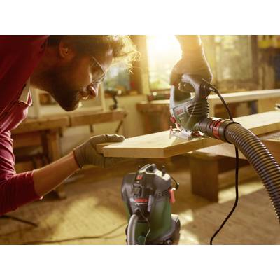 A man is cutting a wooden board with a jigsaw. He is wearing safety glasses and gloves. The workshop is well-lit.