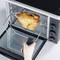 A roasted chicken leg is rotating in an electric grill. A hand is holding the handle of the rotisserie device.